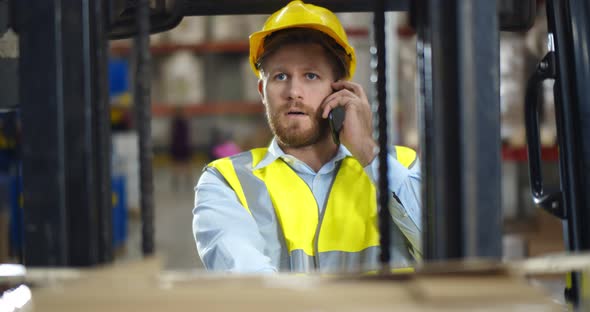 Industrial Warehouse Manager Sitting in Forklift Truck and Having Phone Call
