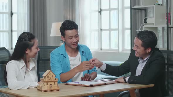 Asian Real Estate Agent Giving The Keys To A Couple After Signing On House Purchase Contract Paper