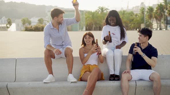 Smiling Diverse Friends Clinking Bottles While Drinking Beer at Sunset on Seafront During Weekend