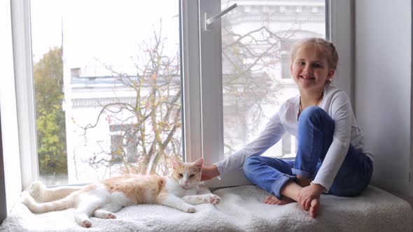 A Little Girl is Stroking Her Cat Sitting on the Windowsill