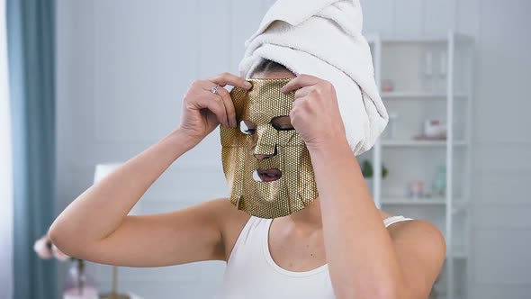 Young Caucasian Woman Applying Rejuvenating Cosmetic Golden Tissue Mask on Face.