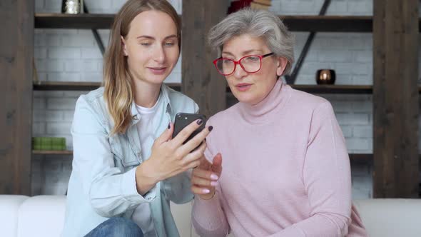 Elderly Mother and an Adult Daughter Are Sitting on the Couch at Home, Having Fun Using a Smartphone