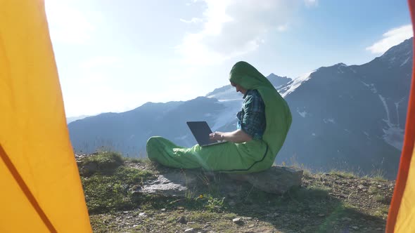 Man in Sleeping Bag Types on Laptop Sitting on Hill Top