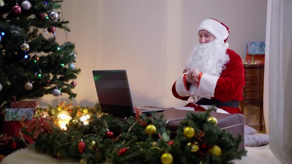 Confident Santa Clause Sitting in Decorated Living Room Indoors Typing on Laptop Keyboard