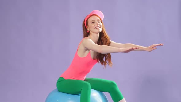 Female Aerobics Trainer in Perfect Shape Sits on Fitness Ball with Hands Forward