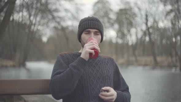 Portrait of Handsome Man Sitting on the Bench Near the River in the Spring Park Drinking Tea