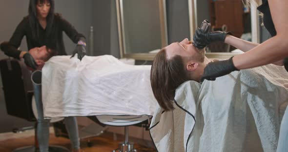 Young Bearded Man is Sitting on the Barber's Chair with Cape Facing the Mirror Female Barber Cuts