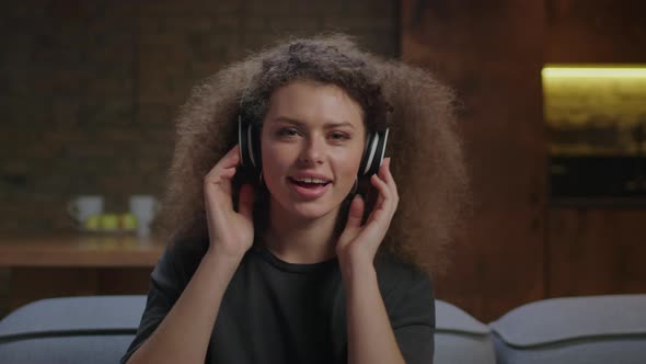 20s Young Woman Enjoys Listening Music in Wireless Headphones Sitting on Couch