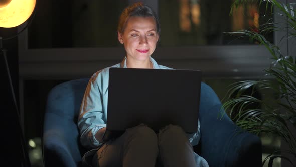 Woman is Sitting in the Armchair and Working on a Laptop at Night