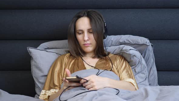 Sleepy young woman in night underwear lying in bed and listening to music in headphones