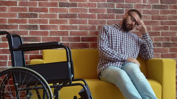 Thoughtful Bearded Disabled Man with Glasses is Sitting Next to a Wheelchair on Sofa Sad and