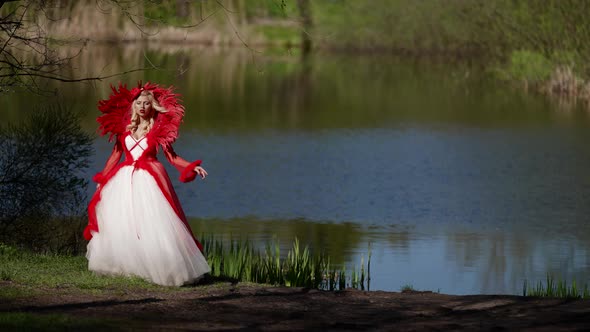 Fairytale Shot with Red Queen or Sorceress Woman in Red Dress on Shore of Calm Lake in Forest