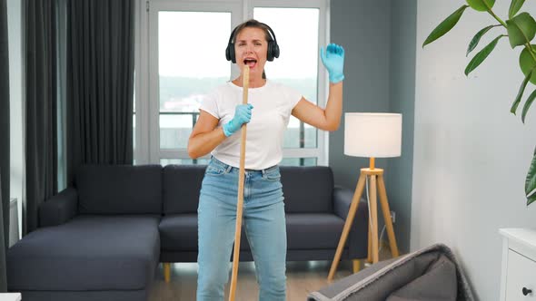 Woman in Headphones Cleans the House and Have Fun Singing with a Broom Like a Star at a Concert