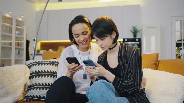 Two Young Girls Look at Their Smartphones and Have Fun Sitting on the Couch at Home