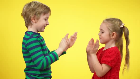 Side View of Excited Carefree Boy and Girl Playing Hand Clapping Game at Yellow Background