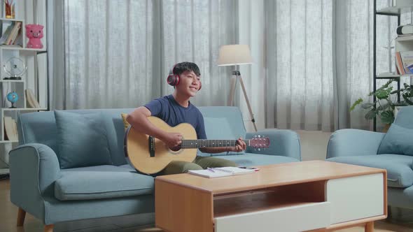 Asian Boy Composer With Notebook On Table Wearing Headphones And Playing Guitar At Home