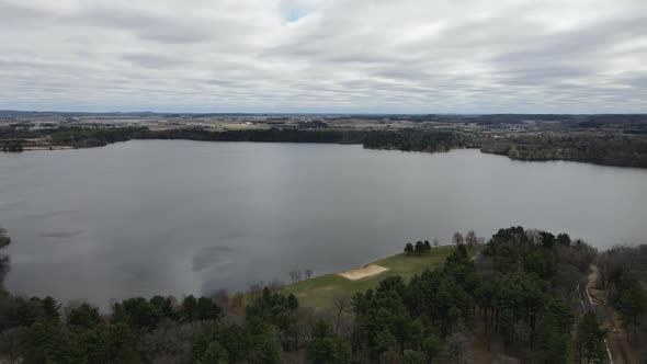 View of lake on a gray cloudy day, light ripples seen on the water surface. Small park with beach. alt