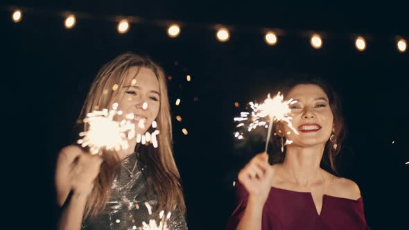 Two Multiracial Girlfriends in Plush Dresses Dancing in Dark at Party Holding and Waving Sparklers