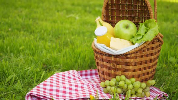 Wicker Picnic Basket with Healthy Food on Red Checkered Table Cloth on Green Grass Outside in Summer