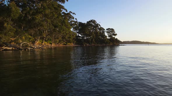 Revealing drone view travelling over calm water next to a rugged coastline covered in native bushlan