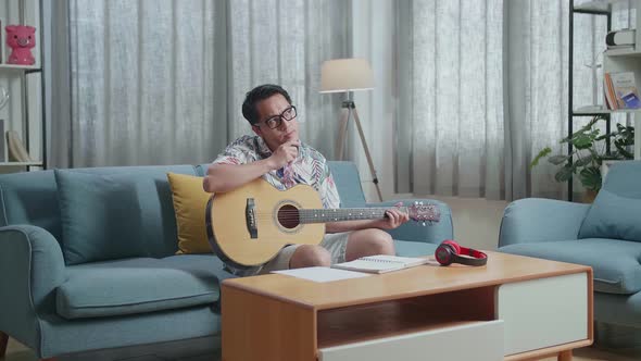 Asian Man Composer With A Guitar Thinking Before Composing Music On Paper At Home