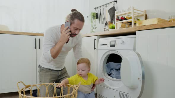 Caucasian busy father use phone call for work and doing housework with baby boy toddler in kitchen.