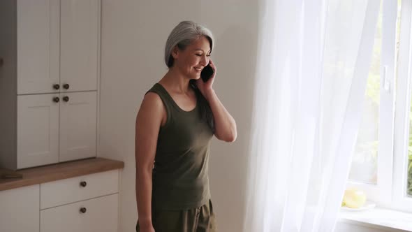 Positive Asian woman talking by phone and looking at window