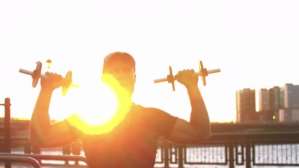 Young Sportive Man Training His Hands with Dumbbells Outdoors on Sunset