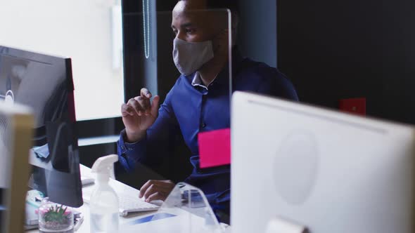 African american man wearing face mask using computer while sitting on his desk at modern office