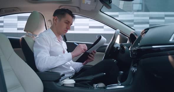 Portrait of Serious Male Entrepreneur Sits on Driver's Seat in Car Reads and Signs Paper Documents