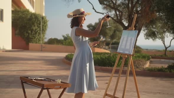 Side View of Smiling Caucasian Slim Woman Shaking Red Wine in Glass Standing at Easel with Picture