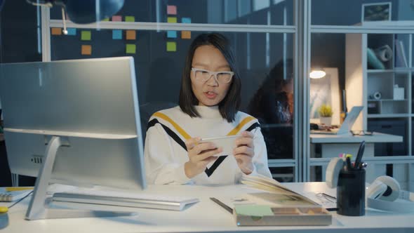 Happy Asian Girl Relaxing in Office Playing Mobile Video Game Touching Smartphone Screen at Night