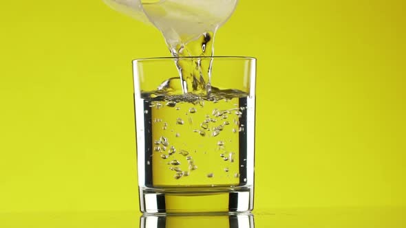 Water is Pouring Down Into Clear Glass Standing on Yellow Background in Slowmotion