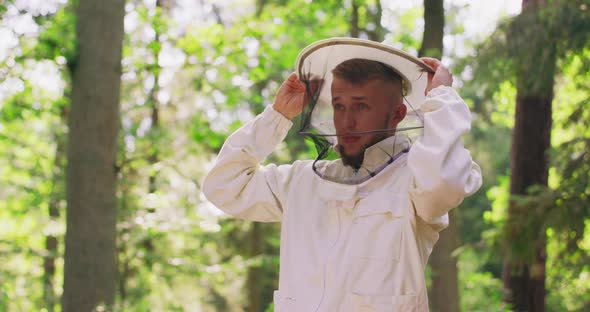Male Bearded Young Handsome Beekeeper Fastens a Zipper of White Protective Suit and Wears a Round