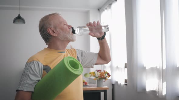 Handsome Elderly Man with Beard After Playing Sports Holds a Yoga Mat in His Hands and Drinks Clean
