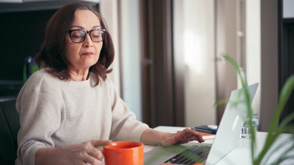 A Senior Woman in Glasses Working at Home with a Laptop and Drinking Coffee