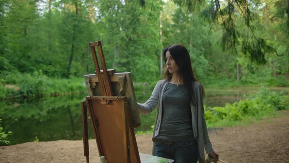Woman Artist in a Blouse and Jeans Paints a Landscape in a Park Near a Pond