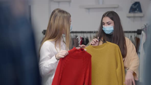 Charming Confident Caucasian Women in Covid Face Masks Changing Hangers with Red and Yellow Sweaters