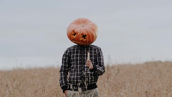 A Man in a Giant Pumpkin Head Stands with Candles in His Hands in the Middle of a Field on a