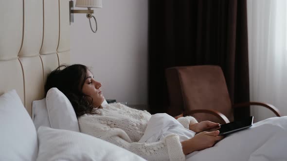 Sleepy Woman Lying on Bed with Tablet in Hands in Modern Apartment