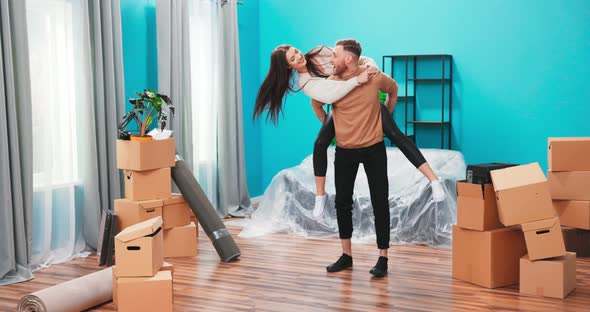 Happy Young Husband Lifting Excited Wife Celebrating Moving Day with Cardboard Boxes Proud Overjoyed