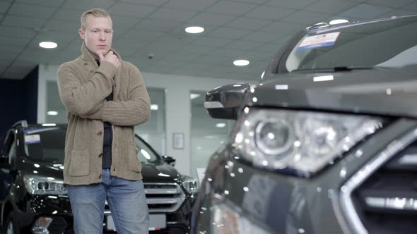 Portrait of Thoughtful Young Caucasian Man Choosing Automobile in Dealership or Showroom, Serious