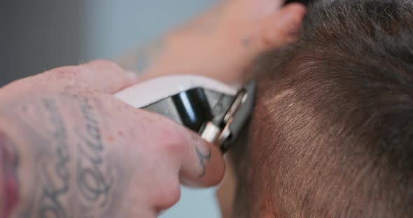 Closeup Tattooed Male Barber's Hands with Cliper is Clippering Hair of Young Man Who is Sitting on