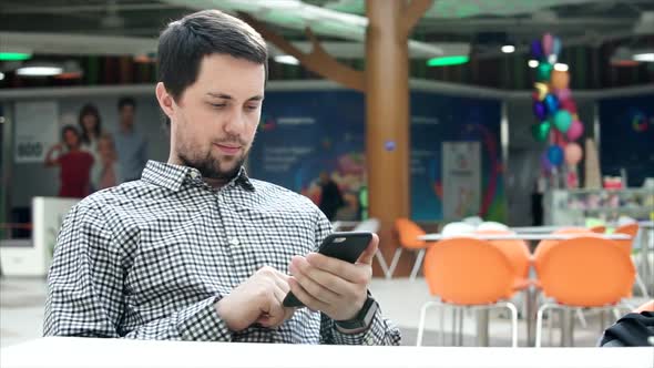 A Young Man Looks at Photos on a Mobile Phone in a Shopping Center Mall.