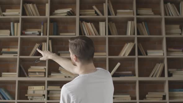 Back View of Brunette Man Searching Book on Shelves in Library. Young Caucasian Guy with Eyeglasses