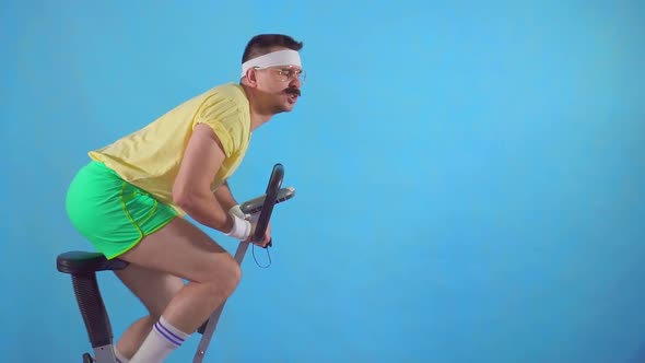 Funny Young Man From the 80's with a Mustache and Glasses on Exercise Bike on a Blue Background Slow