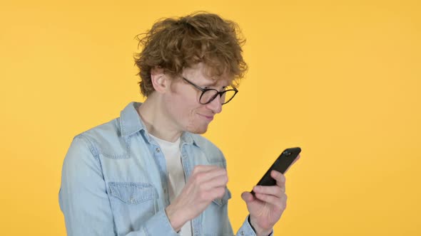 Redhead Young Man Celebrating on Smartphone Yellow Background