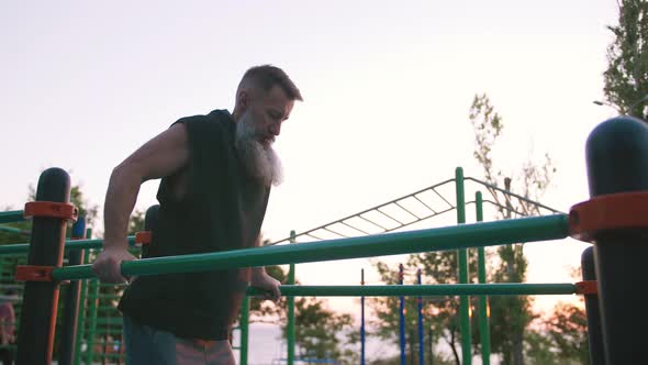 Fit Handsome Middle Aged Man with Long Gray Beard Doing Push Ups on Parallel Bars on Sports Ground