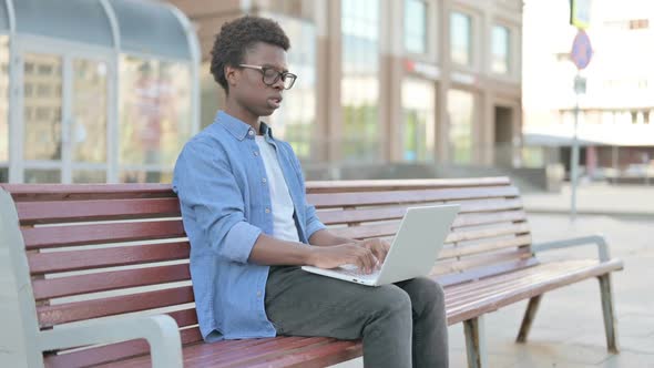 Young African Man with Wrist Pain Using Laptop While Sitting on Bench