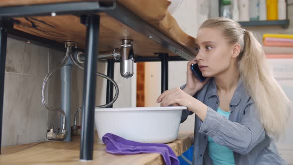 Woman Sitting Near Leaking Sink in Laundry Room Calling for Help